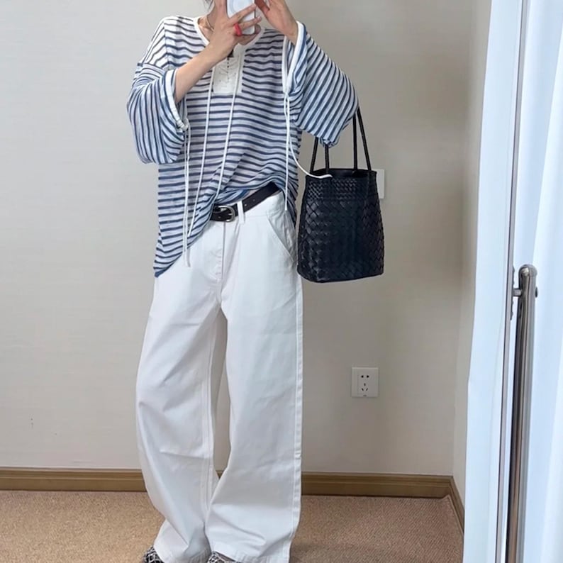 Puede incluir: Un atuendo casual que presenta una parte superior a rayas azules y blancas con mangas largas y cordones blancos. El atuendo incluye pantalones blancos de pierna ancha, un cintur&oacute;n negro y un bolso tote negro tejido. El atuendo se completa con zapatos estampados.