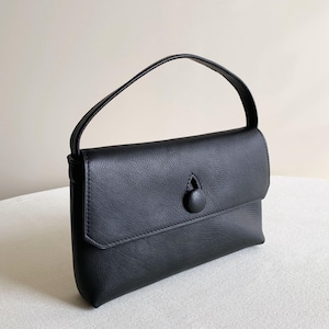May include: A black handbag with a textured surface, featuring a single top handle and a front flap closure secured by a central circular button. The bag has a rectangular shape with stitched edges and a clean, structured design for daily use.