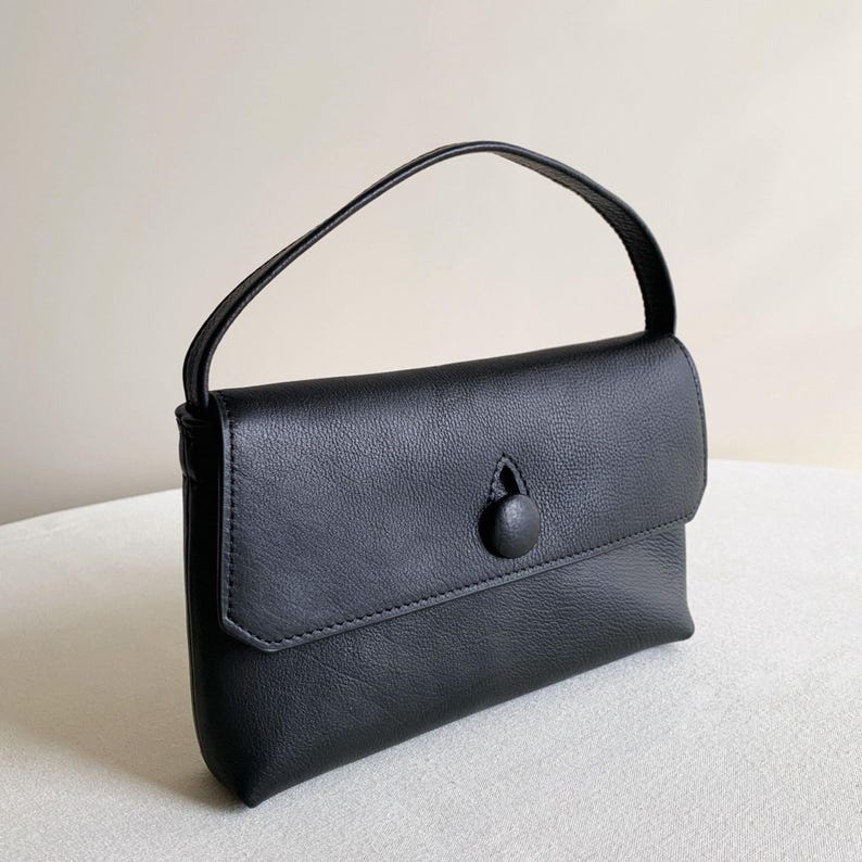 May include: A black handbag with a textured surface, featuring a single top handle and a front flap closure secured by a central circular button. The bag has a rectangular shape with stitched edges and a clean, structured design for daily use.