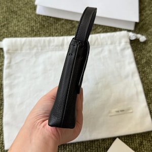 May include: A black leather pouch with a slim, rectangular profile and a single top handle. The exterior features a textured grain finish with visible stitching along the edges. The design is minimalist, focusing on the vertical orientation of the bag.