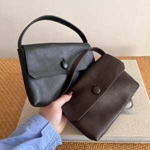 May include: Two structured handbags with a pebbled texture, one in black and one in dark brown. Each bag features a single top handle, a fold-over flap closure, and a large, circular, matching-colored button centered on the front flap.