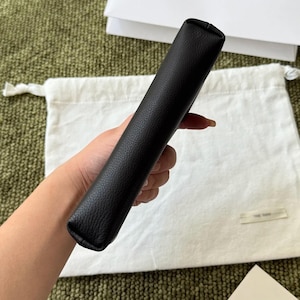 May include: A black rectangular pouch with a textured surface. The item features a slim, elongated shape with rounded edges and a smooth, consistent finish. The pouch is held in a vertical orientation, showcasing its side profile and construction.