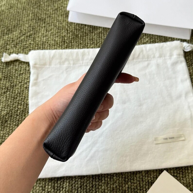 May include: A black rectangular pouch with a textured surface. The item features a slim, elongated shape with rounded edges and a smooth, consistent finish. The pouch is held in a vertical orientation, showcasing its side profile and construction.