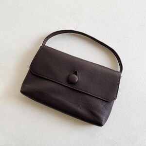 May include: A dark brown handbag with a textured surface, featuring a single top handle and a front flap closure secured by a matching circular button. The bag has a rectangular shape with rounded corners and a clean, simple design.