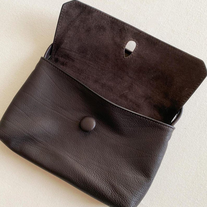 May include: A dark brown leather clutch bag with a pebbled exterior texture and a smooth suede interior flap. The bag features a single circular button closure on the front and a teardrop-shaped cutout on the underside of the top flap.