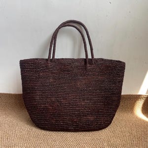 May include: A dark brown woven tote bag with two handles. The bag has a rectangular shape with rounded corners and a textured surface. The handles are made of the same woven material. The bag is sitting on a light brown surface.