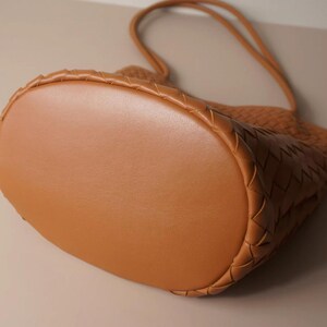 Puede incluir: Un bolso tote de cuero marr&oacute;n con un dise&ntilde;o tejido. El bolso tiene una base redondeada y un asa &uacute;nica. El cuero parece liso y el bolso es de un color c&aacute;lido y neutro.