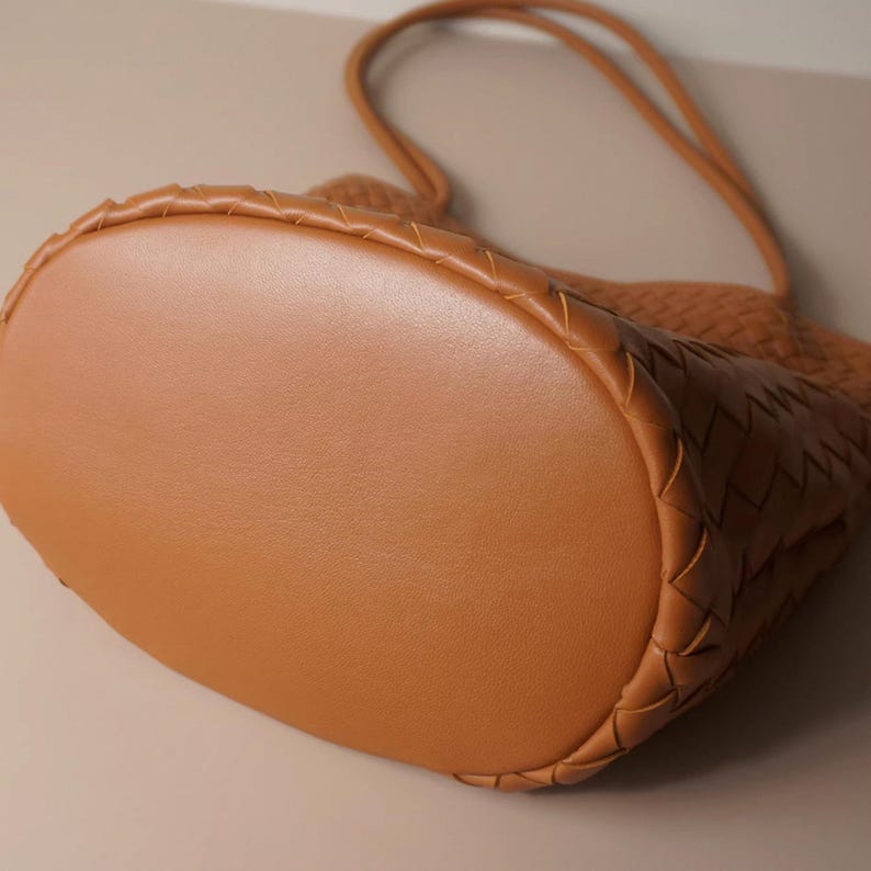 Puede incluir: Un bolso tote de cuero marr&oacute;n con un dise&ntilde;o tejido. El bolso tiene una base redondeada y un asa &uacute;nica. El cuero parece liso y el bolso es de un color c&aacute;lido y neutro.