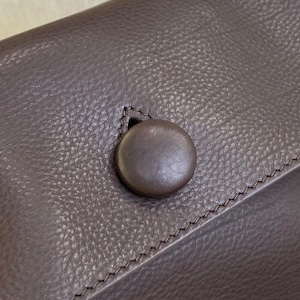 May include: A dark brown textured surface featuring a single circular button closure. The button is smooth and matches the color of the surrounding material, which displays a distinct pebbled grain pattern and visible stitching along the edge.