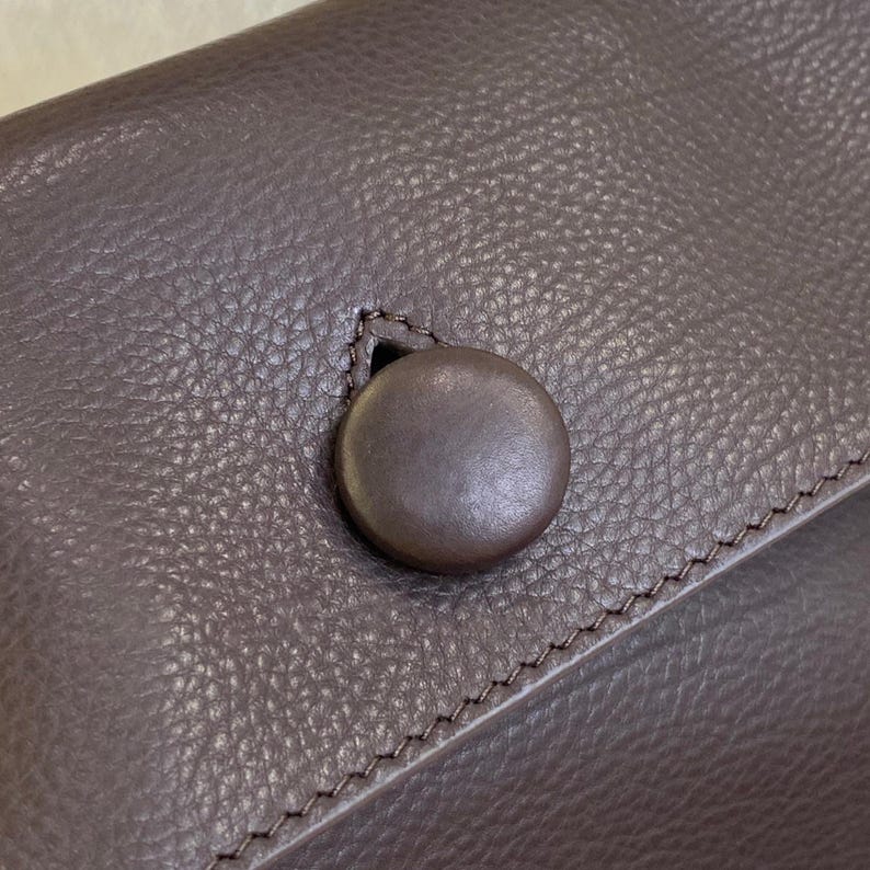 May include: A dark brown textured surface featuring a single circular button closure. The button is smooth and matches the color of the surrounding material, which displays a distinct pebbled grain pattern and visible stitching along the edge.