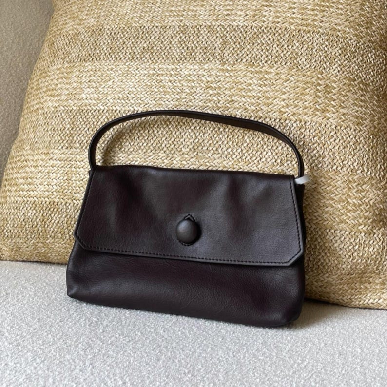 May include: A dark brown rectangular handbag featuring a single top handle and a front flap closure secured by a matching circular button. The bag has a smooth, matte finish and a structured silhouette with clean, minimalist stitching along the edges.