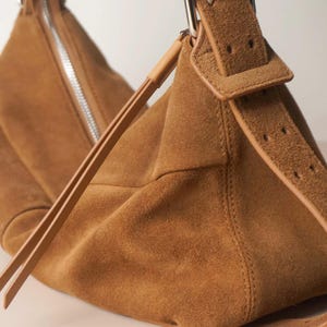 May include: A tan suede shoulder bag with a silver zipper and a matching adjustable strap. The bag features a rectangular buckle and a soft, textured surface. The bag is a neutral color and has a minimalist design.
