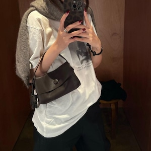 May include: A dark brown leather handbag with a single top handle and a circular button closure, paired with a white short-sleeved t-shirt featuring a black graphic print, a grey knit sweater draped over the shoulders, and black trousers.
