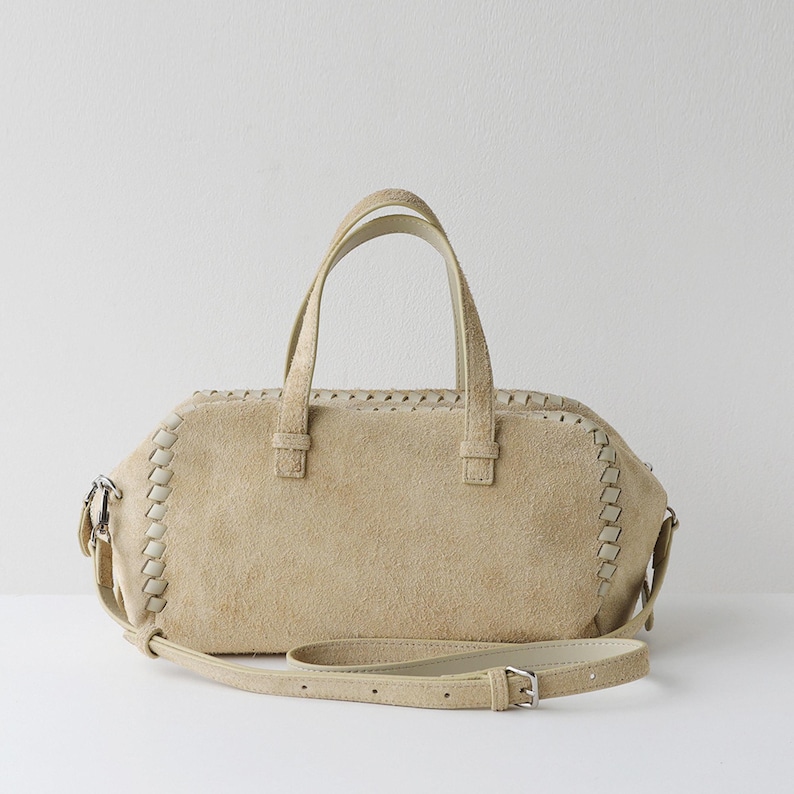Puede incluir: Bolso de mano de ante beige con asa superior y bandolera extra&iacute;ble. El bolso presenta costuras decorativas en los bordes y herrajes plateados. El bolso mide aproximadamente 30 cm de ancho.