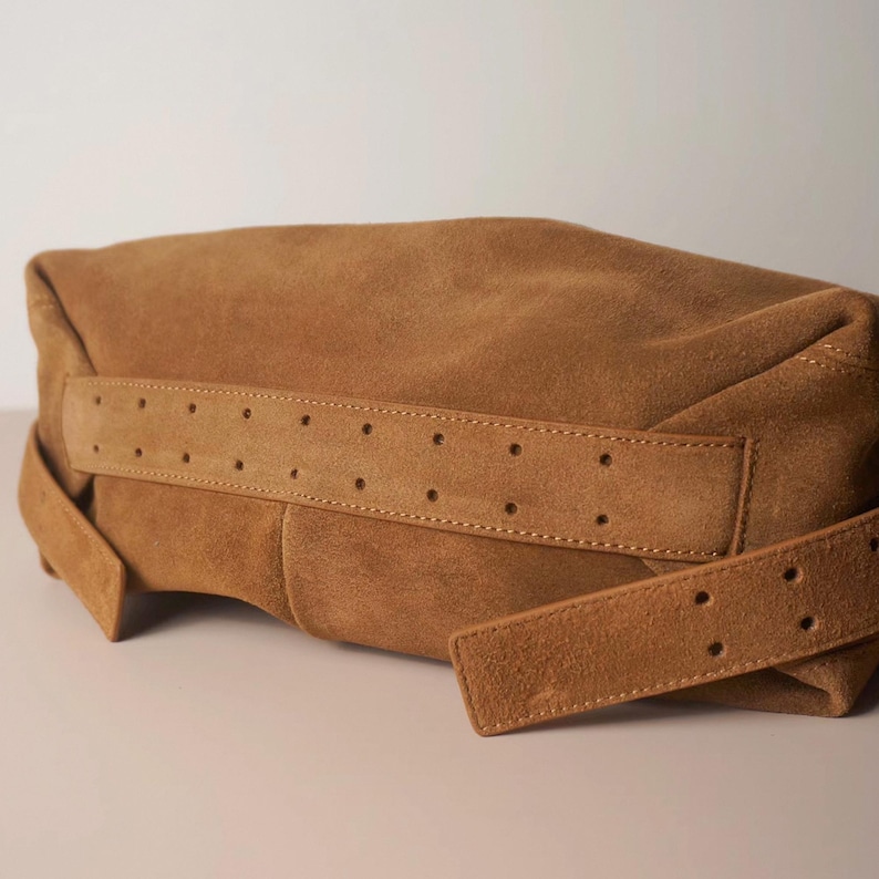May include: A tan suede clutch bag with a wide, perforated strap detail. The bag has a soft, slouchy shape and a matching strap that wraps around the body of the bag. The color is a warm, earthy tone.