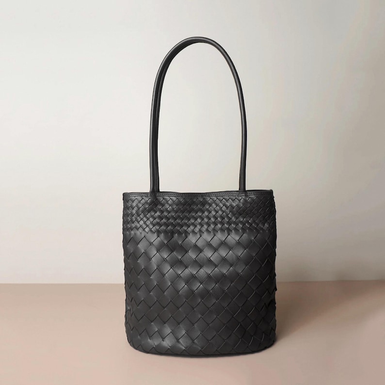 Puede incluir: Bolso tote negro con dise&ntilde;o tejido. El bolso tiene un cuerpo rectangular con base redondeada y dos asas largas. Las asas est&aacute;n unidas a la parte superior del bolso. El bolso est&aacute; hecho de un material oscuro.