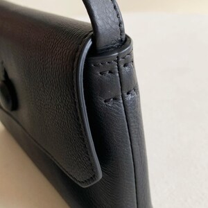 May include: A black leather handbag featuring a textured grain surface, a flap closure with a single button, and a strap attachment point on the side. The bag is constructed with visible stitching along the edges and a smooth, rounded side profile.