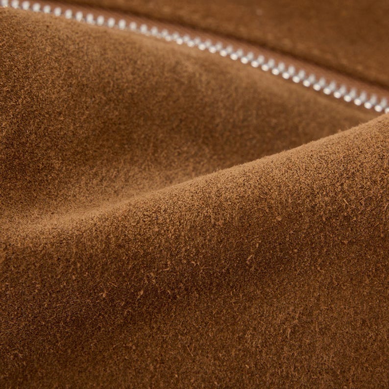 May include: Close-up of brown suede fabric with a silver zipper. The suede has a soft, textured appearance with subtle variations in color. The zipper is visible along the top edge, adding a contrasting element to the design.