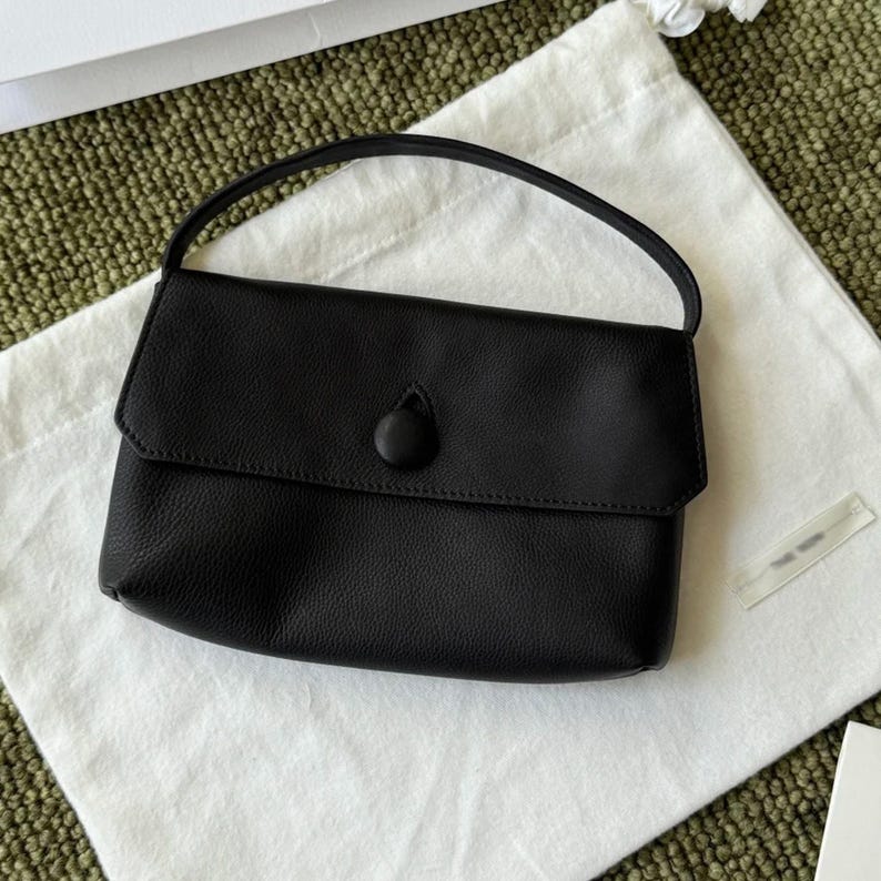 May include: A black leather handbag with a single top handle and a fold-over flap closure. The flap is secured by a single, centered, fabric-covered button. The bag features a pebbled texture and a structured, rectangular silhouette.