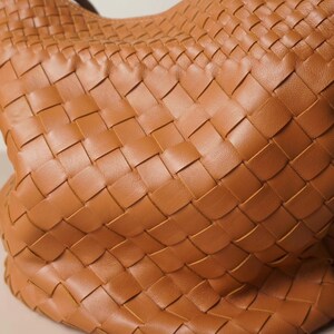 Puede incluir: Primer plano de un bolso de mano de cuero tejido en color marr&oacute;n c&aacute;lido. La textura del bolso se crea mediante un patr&oacute;n repetitivo de tiras entrelazadas, lo que le da un aspecto &uacute;nico y elegante. El dise&ntilde;o sugiere un interior espacioso.