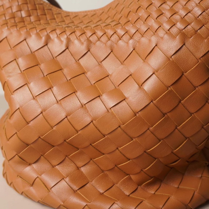 Puede incluir: Primer plano de un bolso de mano de cuero tejido en color marr&oacute;n c&aacute;lido. La textura del bolso se crea mediante un patr&oacute;n repetitivo de tiras entrelazadas, lo que le da un aspecto &uacute;nico y elegante. El dise&ntilde;o sugiere un interior espacioso.