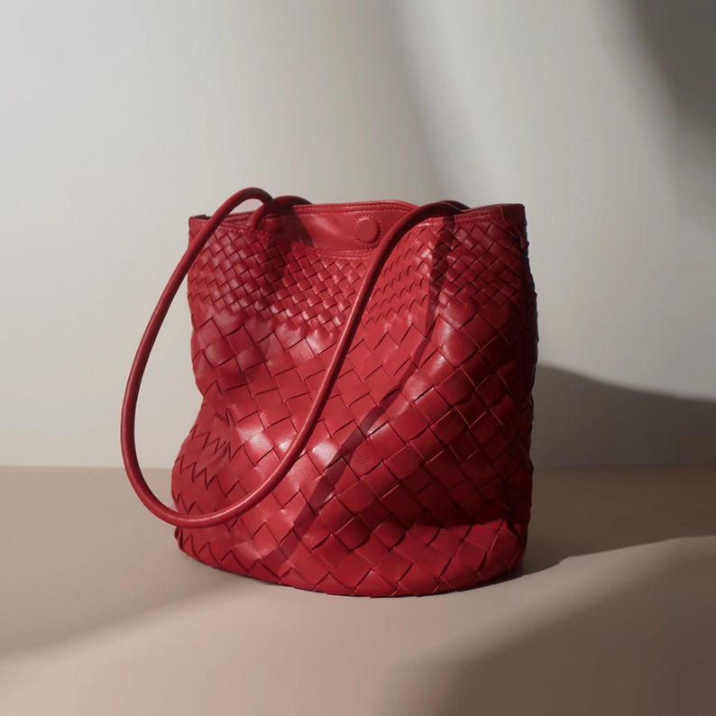 Puede incluir: Un bolso tote de cuero rojo tejido con cierre de bot&oacute;n. El bolso tiene dos asas largas y redondeadas. El bolso est&aacute; hecho de un material de cuero tejido y es de color rojo brillante.