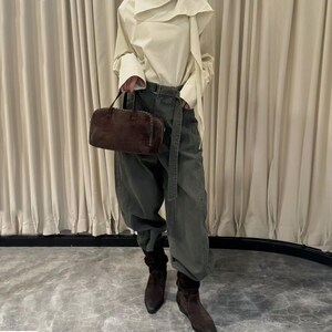 Puede incluir: Una persona con una blusa color crema con cuello drapeado, pantalones verde oliva y botas de ante marrones. Sostiene un bolso de mano de ante marr&oacute;n con asa superior. El atuendo se complementa con un cintur&oacute;n.