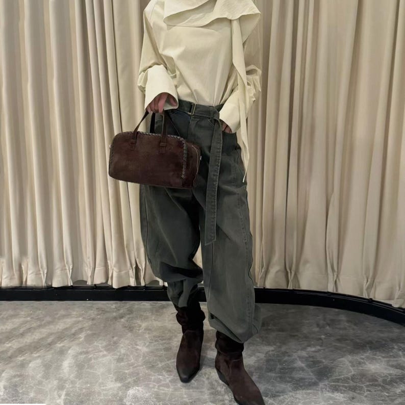 Puede incluir: Una persona con una blusa color crema con cuello drapeado, pantalones verde oliva y botas de ante marrones. Sostiene un bolso de mano de ante marr&oacute;n con asa superior. El atuendo se complementa con un cintur&oacute;n.