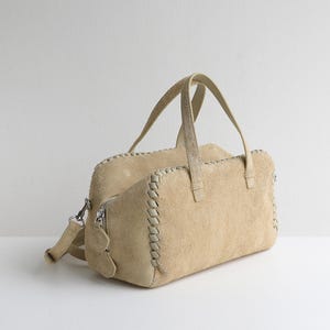Puede incluir: Bolso de mano de ante color beige con forma rectangular. El bolso tiene dos asas y una correa para el hombro desmontable. Los bordes est&aacute;n detallados con un dise&ntilde;o cosido de color crema. Una cremallera y herrajes plateados completan el dise&ntilde;o.