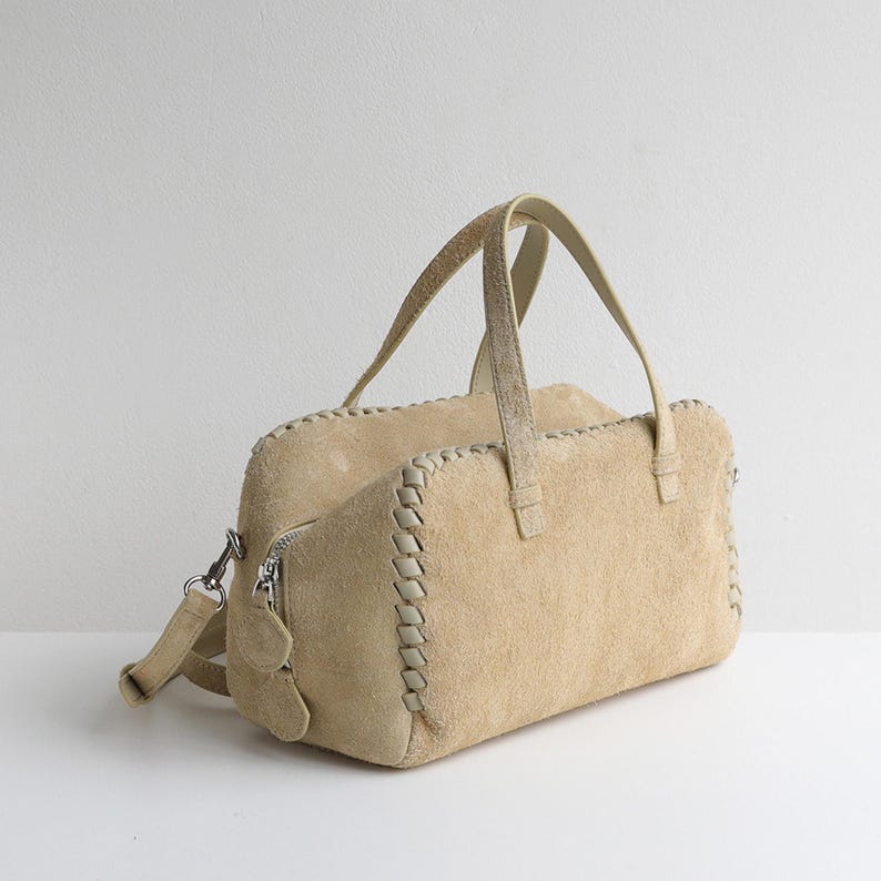Puede incluir: Bolso de mano de ante color beige con forma rectangular. El bolso tiene dos asas y una correa para el hombro desmontable. Los bordes est&aacute;n detallados con un dise&ntilde;o cosido de color crema. Una cremallera y herrajes plateados completan el dise&ntilde;o.