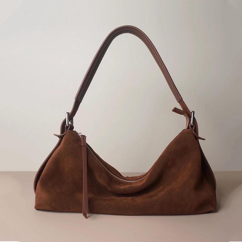 May include: A brown suede shoulder bag with a curved shape and a long, brown leather strap. The bag has a zipper closure and a small, dangling leather pull. The bag is a rich, warm brown color.