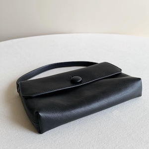 May include: A black leather handbag with a rectangular silhouette, a single top handle, and a front flap closure secured by a large, circular, fabric-covered button. The material features a visible pebbled texture throughout the exterior surface.
