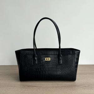 Puede incluir: Un bolso tote negro con un patrón texturizado de relieve de cocodrilo. El bolso cuenta con dos asas finas para el hombro y un cierre giratorio rectangular de metal en tono dorado centrado en el panel frontal. Diseño estructurado.