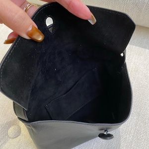 May include: A black leather bag with a smooth exterior and a soft, dark suede interior lining. The bag features a flap closure with a single circular cutout and a small black button fastener located on the front panel, revealing an open compartment.