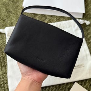 May include: A black leather handbag with a rectangular shape and a single arched top handle. The bag features a pebbled texture and a subtle embossed logo on the lower front center. The design is simple with clean lines and a structured silhouette.