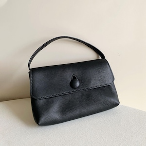 May include: A black handbag with a textured surface, featuring a single top handle and a front flap closure secured by a prominent, circular, fabric-covered button detail centered on the flap. The bag has a structured yet soft silhouette.