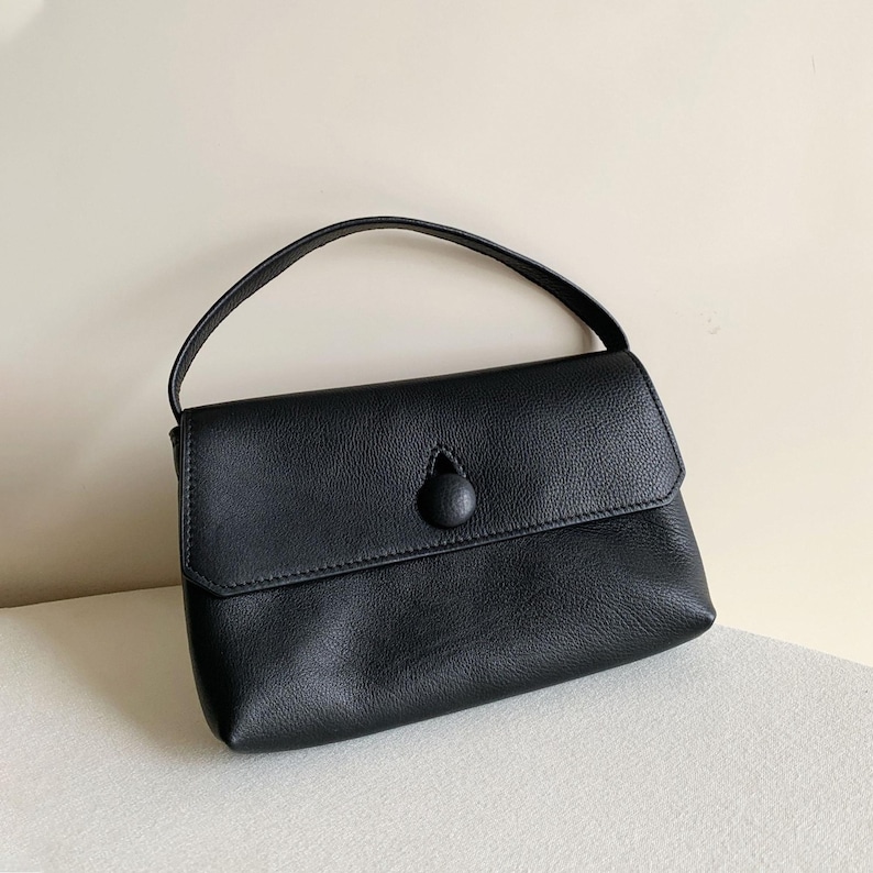 May include: A black handbag with a textured surface, featuring a single top handle and a front flap closure secured by a prominent, circular, fabric-covered button detail centered on the flap. The bag has a structured yet soft silhouette.