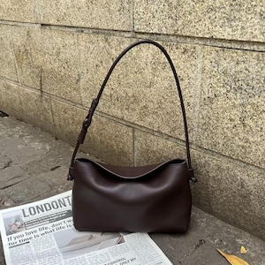 May include: A dark brown shoulder bag with a smooth, matte finish and a curved top opening. The bag features a single, thin, adjustable strap attached to the sides with small knots, creating a simple, structured silhouette for daily use.