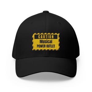 May include: A black baseball cap featuring an embroidered rectangular patch on the front. The patch is yellow with a black border and diagonal stripes, displaying the text CAUTION MUSICAL POWER OUTLET in bold black capital letters.