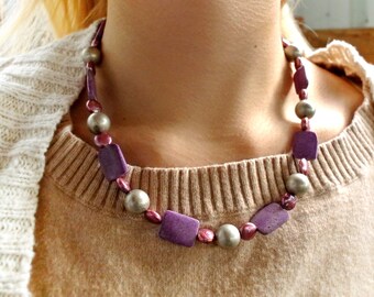 May include: A purple and silver beaded necklace with square and round beads. The necklace is worn by a person with a brown sweater and a white cardigan.