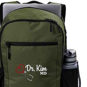 May include: A green backpack with a black zipper and a black water bottle holder. The backpack has a white embroidered design that says "Dr. Kim MD" with a red heart and a stethoscope.