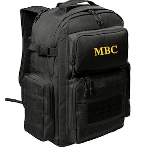 May include: Black tactical backpack with multiple pockets and a large zippered compartment. The backpack has a yellow embroidered logo that says "MBC".