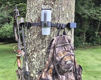 Puede incluir: Un sistema de correas negras ajustables montado alrededor de un tronco de árbol, con un portavasos central con un vaso gris, un gancho lateral que sostiene un arco compuesto con patrones de camuflaje y una mochila de camuflaje con bolsillos.