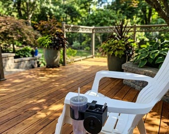 May include: A black plastic cup holder attachment secured to the arm of a white outdoor chair. The accessory features a circular opening holding a clear plastic tumbler with a straw and a rectangular compartment containing a set of metal keys.