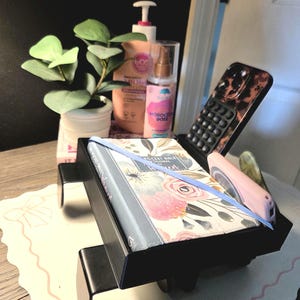 May include: A black bedside organizer holds a floral-covered book with a blue ribbon, a pink pen, and a phone with a tortoiseshell case. A small potted plant and bottles of lotion and spray are also visible.