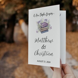 Puede incluir: Una invitación de boda blanca con un diseño floral y una pila de libros. El texto dice "A New Chapter Begins..."  "Matthew & Christina" y "August 21, 2024".
