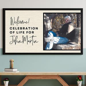 May include: A framed memorial sign with the text "Welcome! Celebration of Life for John Martin" in elegant script. The right side features a photo of a person seated outdoors. The frame is black.