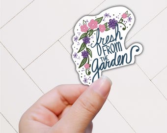 May include: A white sticker with the text "fresh from the garden" in a script font, surrounded by pink and purple flowers and green leaves. The sticker is held by a hand.