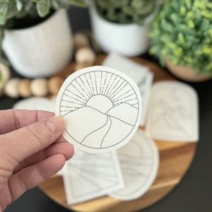 May include: A black and white line drawing of a sun rising over mountains. The sun has rays emanating from it. The drawing is on a white circular sticker.