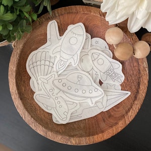 May include: A set of white paper space-themed stickers with outlines of a rocket, spaceship, airplane, hot air balloon, and stars. The stickers are in a wooden bowl.